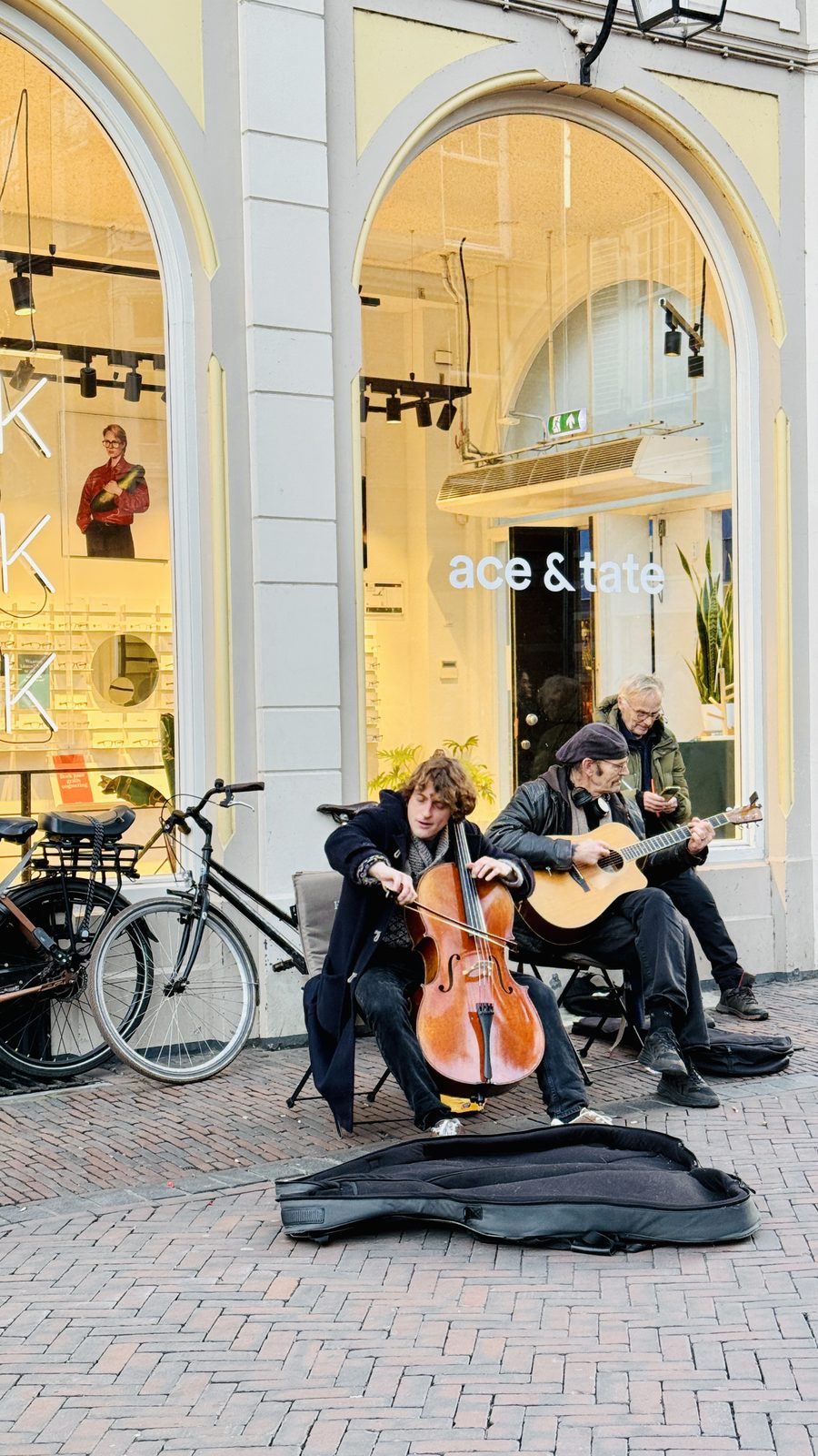 Two street musicians — a cellist and a guitarist — playing outside an Ace & Tate shop in the Netherlands